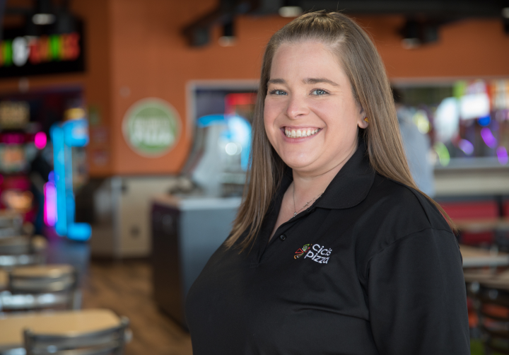 An employee smiles for a portrait in the dining room of a Cicis Pizza. She is wearing a black polo with the Cicis pizza spark logo and text reading 'Cicis Pizza'.