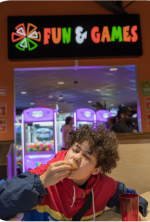 A boy animatedly eats the last few bites of his pizza. Behind him is the Cicis Game Zone with a lit sign over the room featuring the Cicis pizza spark logo and text that reads 'Fun & Games'.