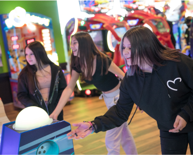 Three teenage girls have fun playing an arcade game together in the Cicis Game Zone. They are next to an arcade machine with a bowling ball graphic on the side.