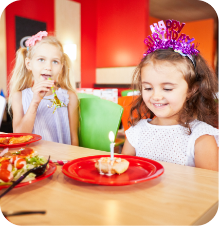 Two young girls sit next to each other at a table in a Cicis Pizza Buffet. One girl is wearing a crown that says "Happy Birthday" and has a Cicis cinnamon roll topped with lit birthday candle in front of her.