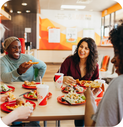 A group of business people smile and chat as they sit together in a Cicis dining room. There is a lit up red "Cicis" sign behind them.