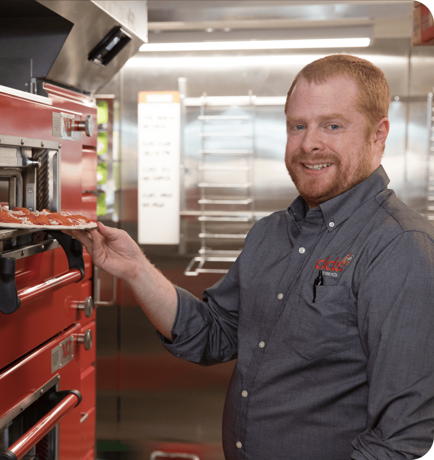 A man wearing a shirt with the Cicis pizza spark logo is putting a pepperoni pizza into red pizza oven.