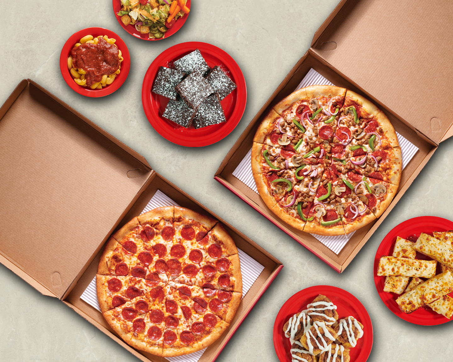 Two pizza boxes, open to show a pepperoni pizza and a supreme pizza. There are four red plates next to the boxes. Pasta and marinara sauce, a stack of brownies, cinnamon rolls, and garlic cheesy bread are plated.