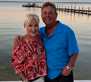 Portrait photo of franchisee Frank & Judy. Smiling couple on a sandy beach at sunset—woman in a red patterned blouse and white shorts, man in a blue shirt and dark shorts.